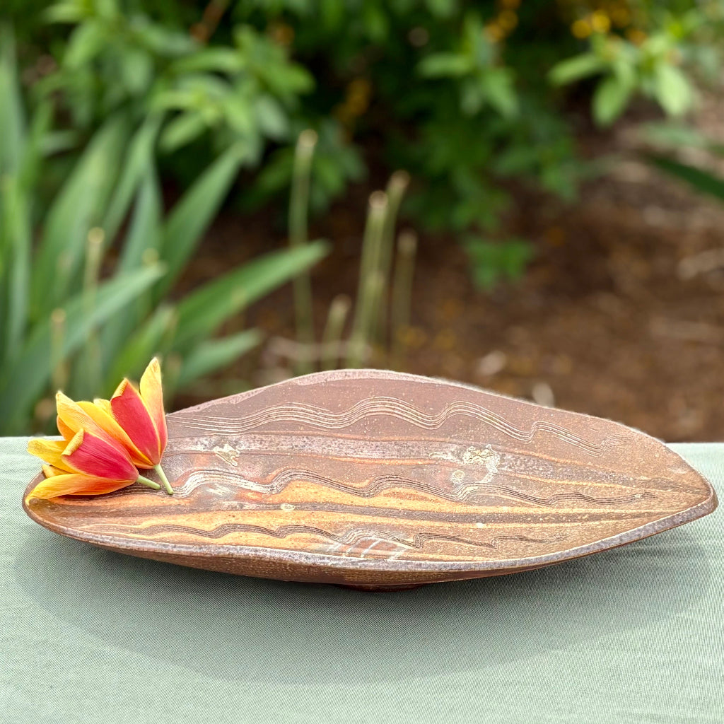 Handmade elongated oval serving tray with natural wave patterns in earthy brown tones on a green cloth with greenery in the background, wood-fired in North Carolina