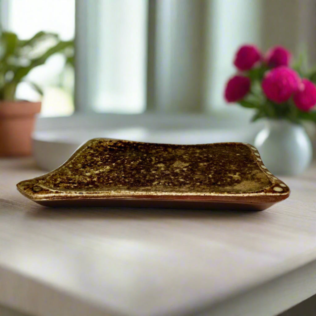 Handmade earth-tone stoneware soap dish with natural speckled texture and slightly raised edges on countertop with flowers and a window in the back.