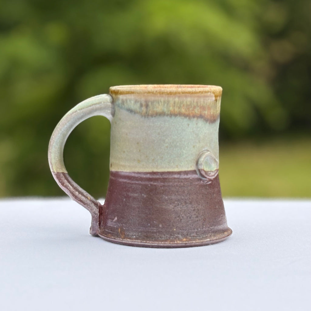 Side View: Pottery mug with an unglazed, earthy lower half and a variegated green upper half. A small button adds visual and tactile interest. Made in Danbury, NC.