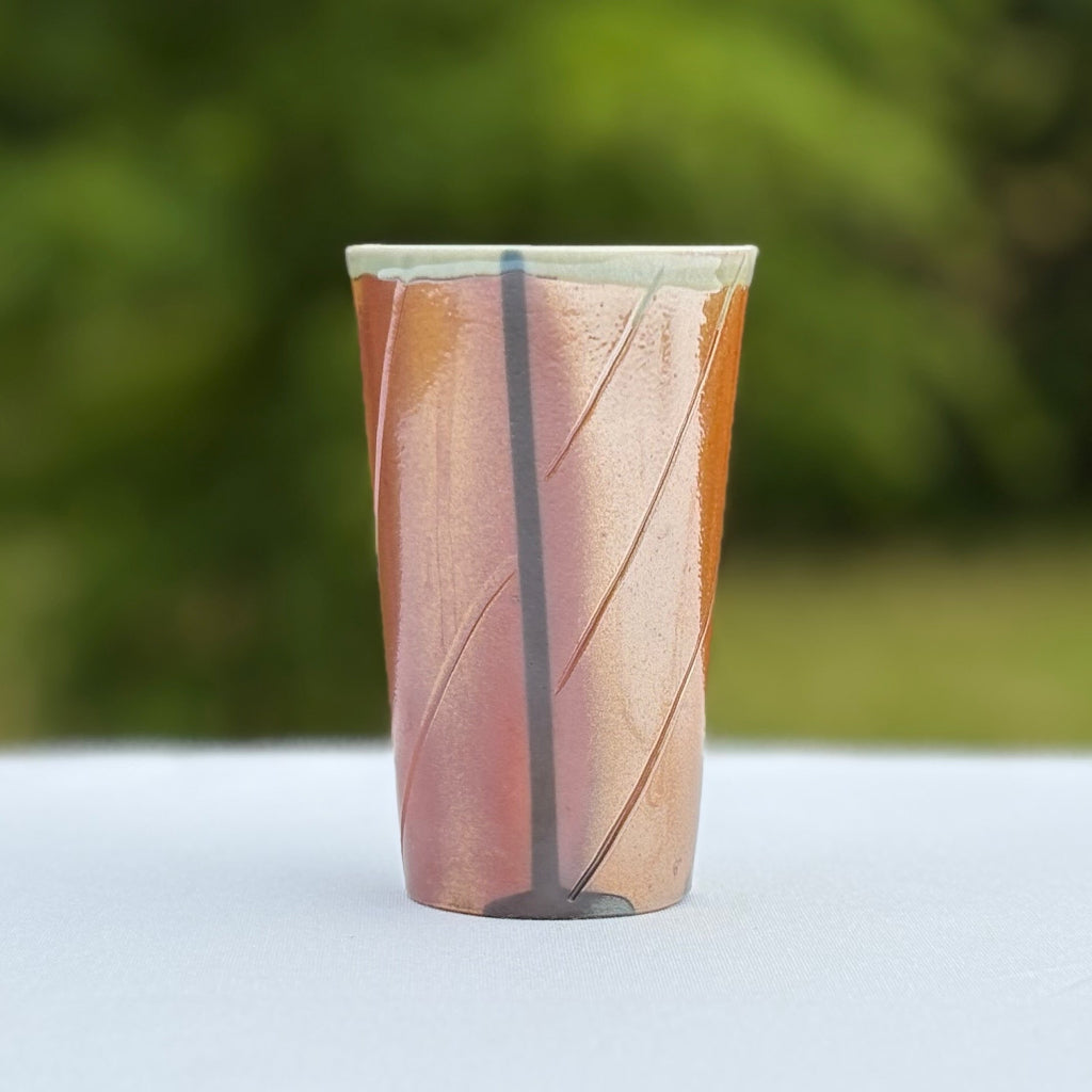 Sideview: Dramatic rich peach porcelain tumbler with a light greenish white glaze, horizontal slashes, an and a vertical black line made in Danbury, NC.
