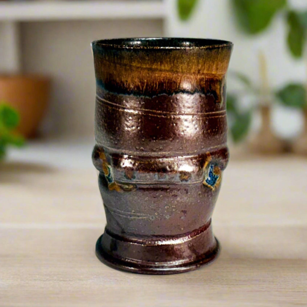 Hand-thrown ceramic tumbler with amber rim gradient and chocolate brown body from wood-firing on a wooden countertop with plants in the background.