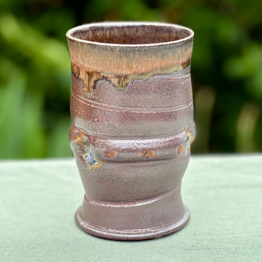 Hand-thrown ceramic tumbler with amber rim gradient and chocolate brown body from wood-firing on a green cloth and greenery in the background. 