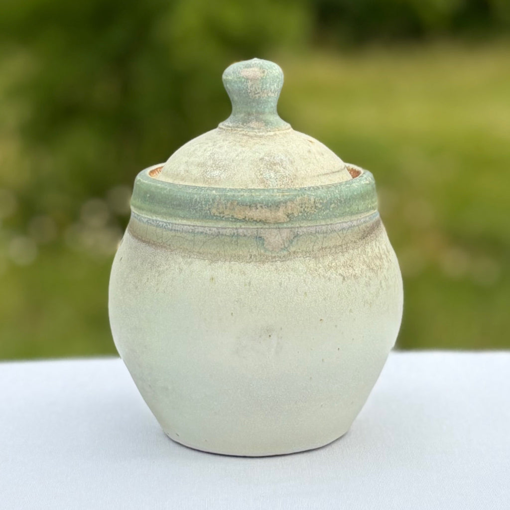 Side View: Lovely round pottery jar and lid with a matte green rim and soft white exterior with delicate lid with a light green knob made in Danbury, NC.
