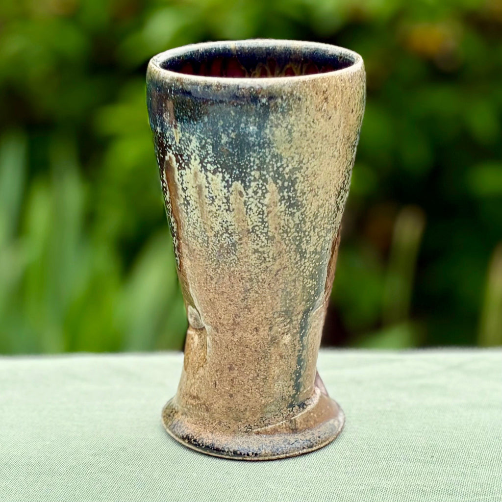 Handcrafted wood-fired ceramic tumbler with amber and natural clay accents on a countertop with greenery in the background.