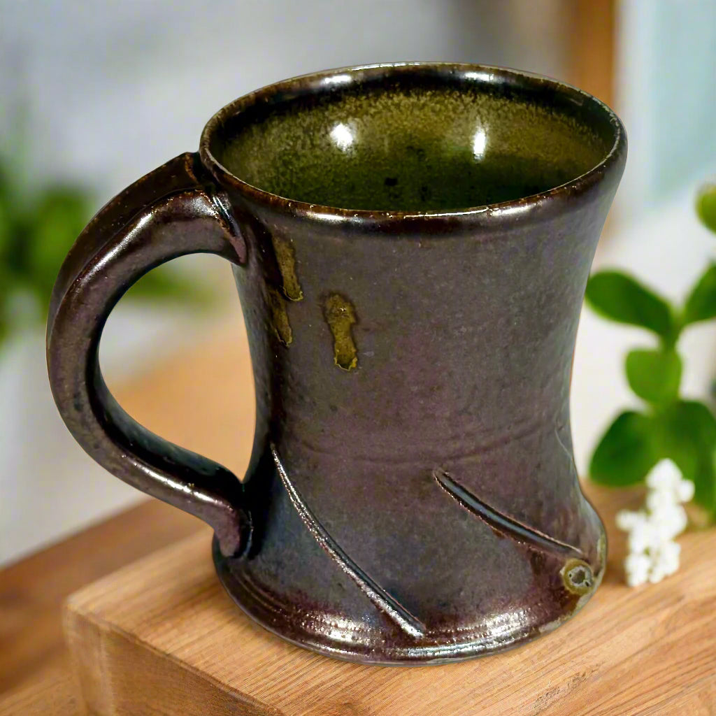 Handcrafted wood-fired ceramic mug with brown finish and decorative keyhole design on counter top with flowers made in North Carolina.