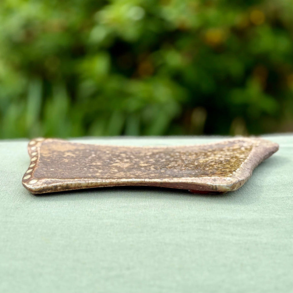 Handmade earth-tone stoneware soap dish with natural speckled texture and slightly raised edges on a green cloth in greenery in the background.