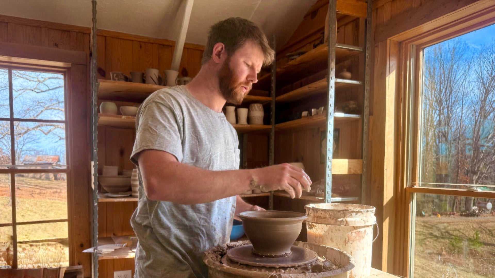 Sam at wheel throwing pot with unfinished pottery on shelves and windows in the background. 