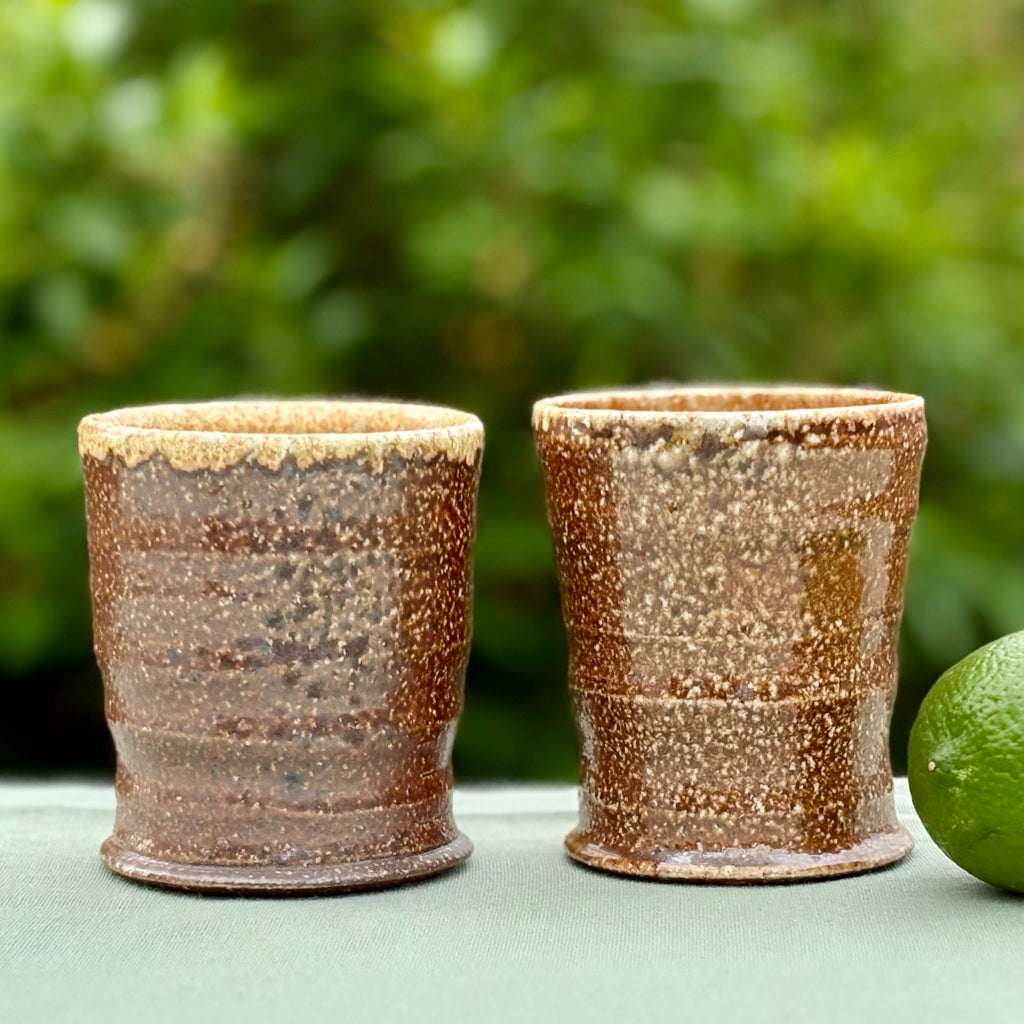 Pair of handcrafted wood-fired ceramic tumblers with sandy textured exterior and warm golden interior glaze on a green cloth with lime and greenery in the background. 