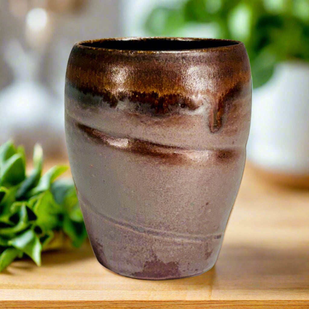 Handcrafted wood-fired ceramic tumbler with mauve-brown body and flowing amber glaze detail on a kitchen counter with herbs in the background.