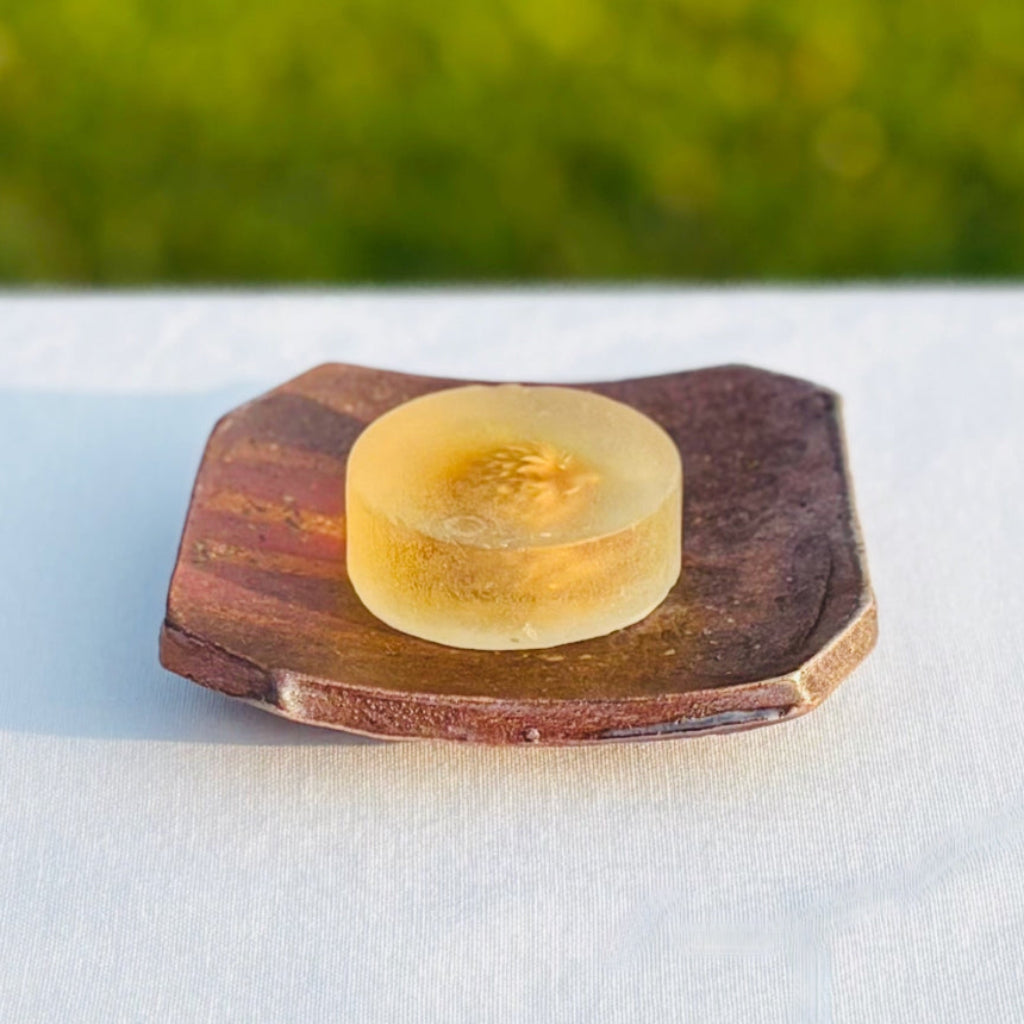 Wood-fired ceramic square soap dish with natural ash glaze showing orange flame patterns and earth tones with three white dots in the center and a subtle lines and a round clear soap in the center. 