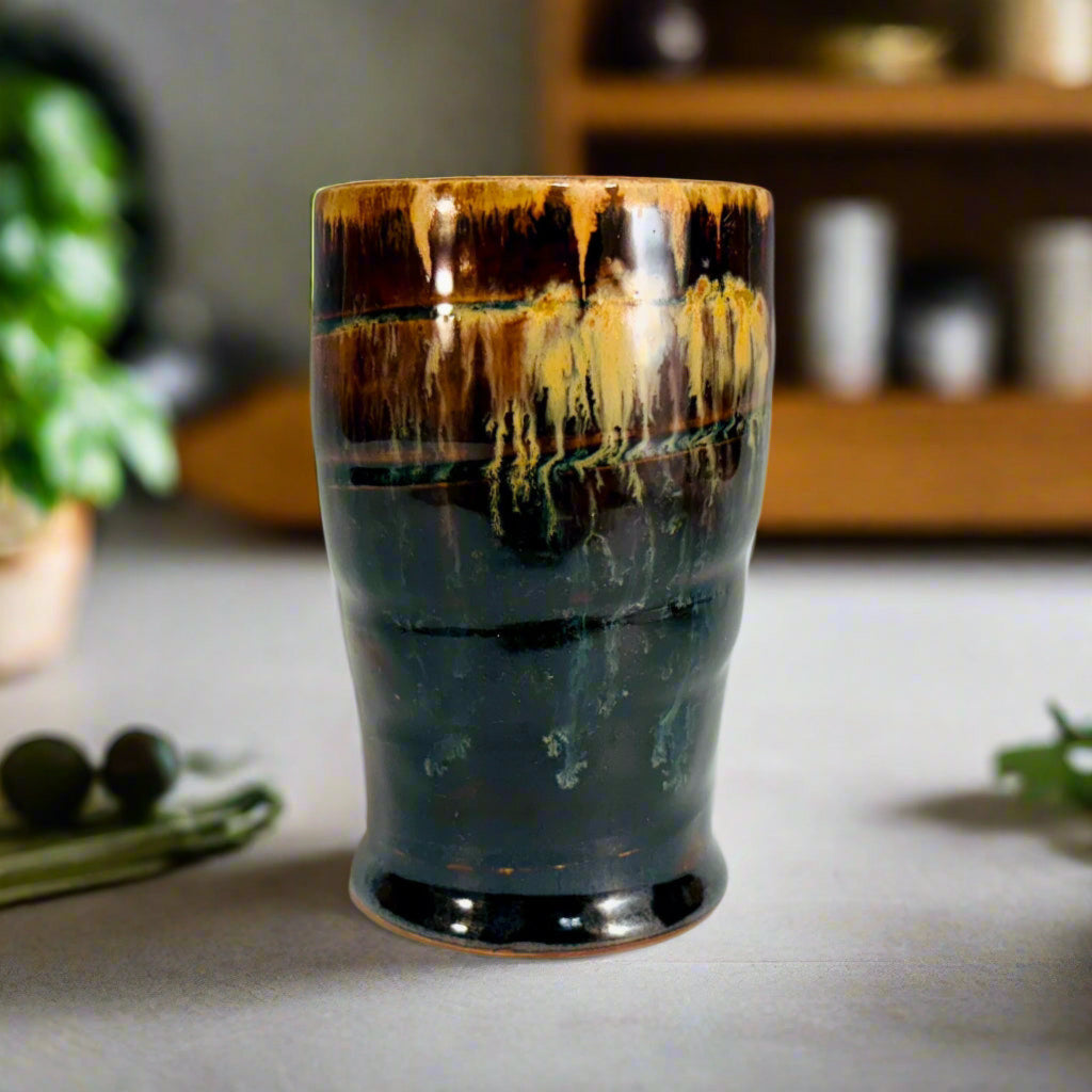 Handcrafted stoneware tumbler with horizontal ridges and blue-black to amber wood-fired glaze effect on a wooden counter top with herbs and a shelves in the background.