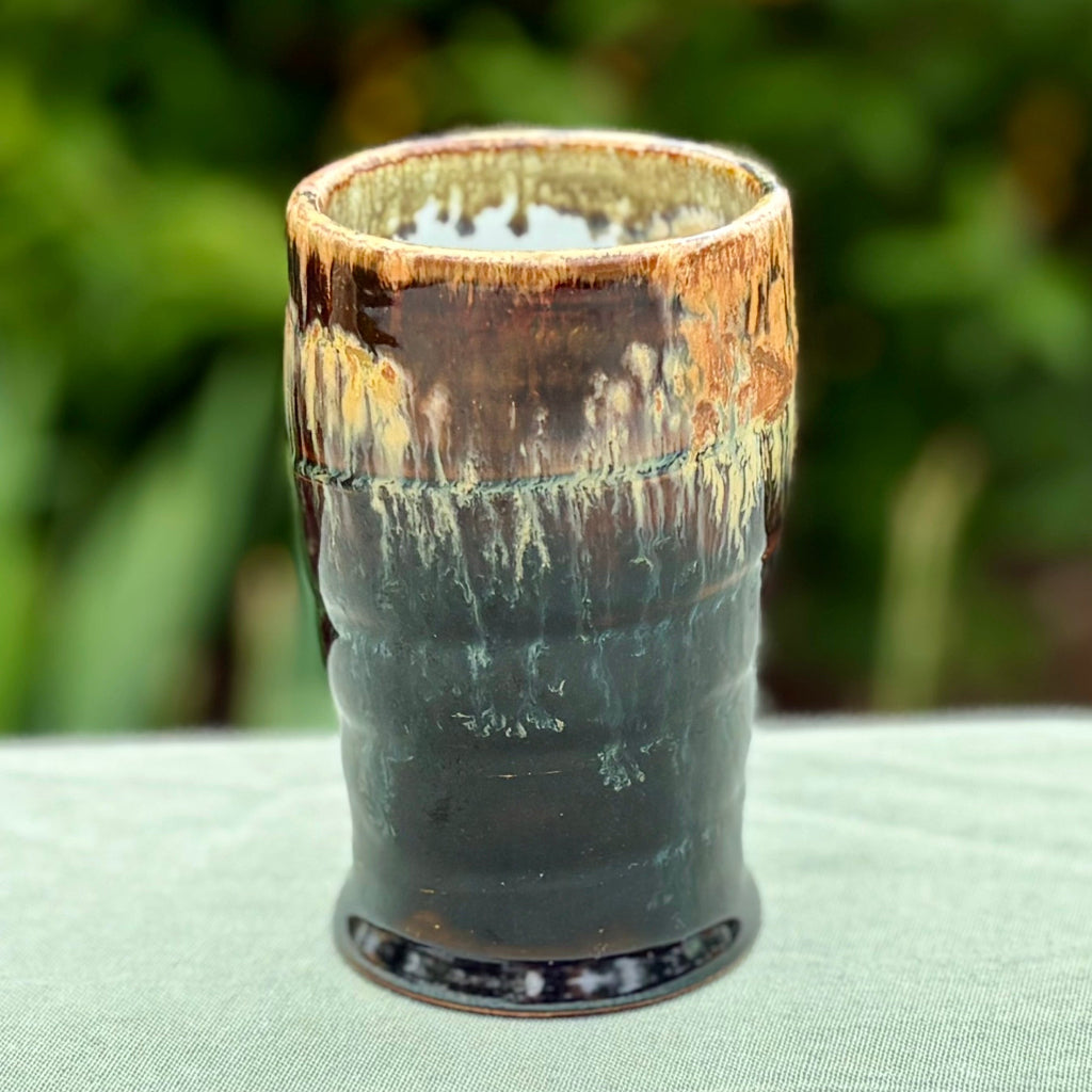 Handcrafted stoneware tumbler with horizontal ridges and blue-black to amber wood-fired glaze effect on a green cloth with greenery in the background. 