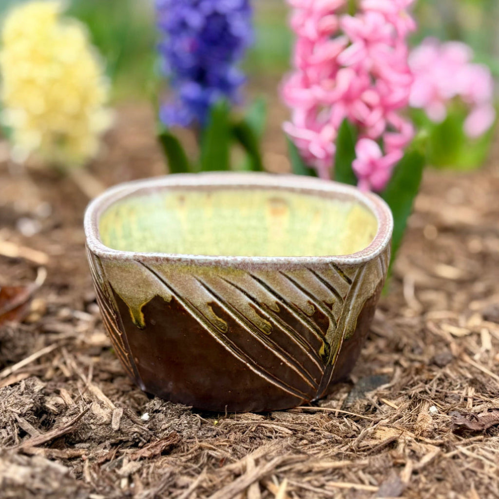 Purple-brown square ceramic planter with hand-carved diagonal pattern and yellow-green interior with flowers in the background.