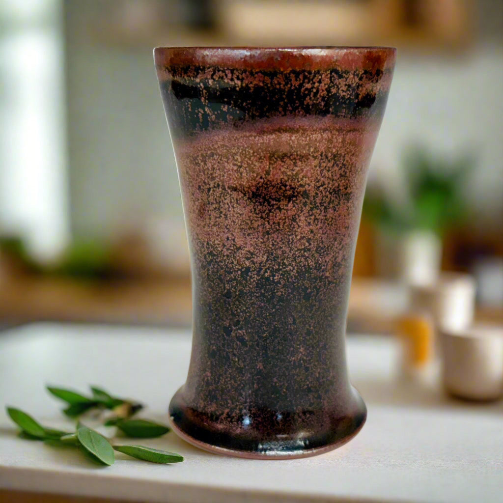 Handcrafted wood-fired cup with red/pink speckles and flared rim on countertop with herbs next to the cup. 