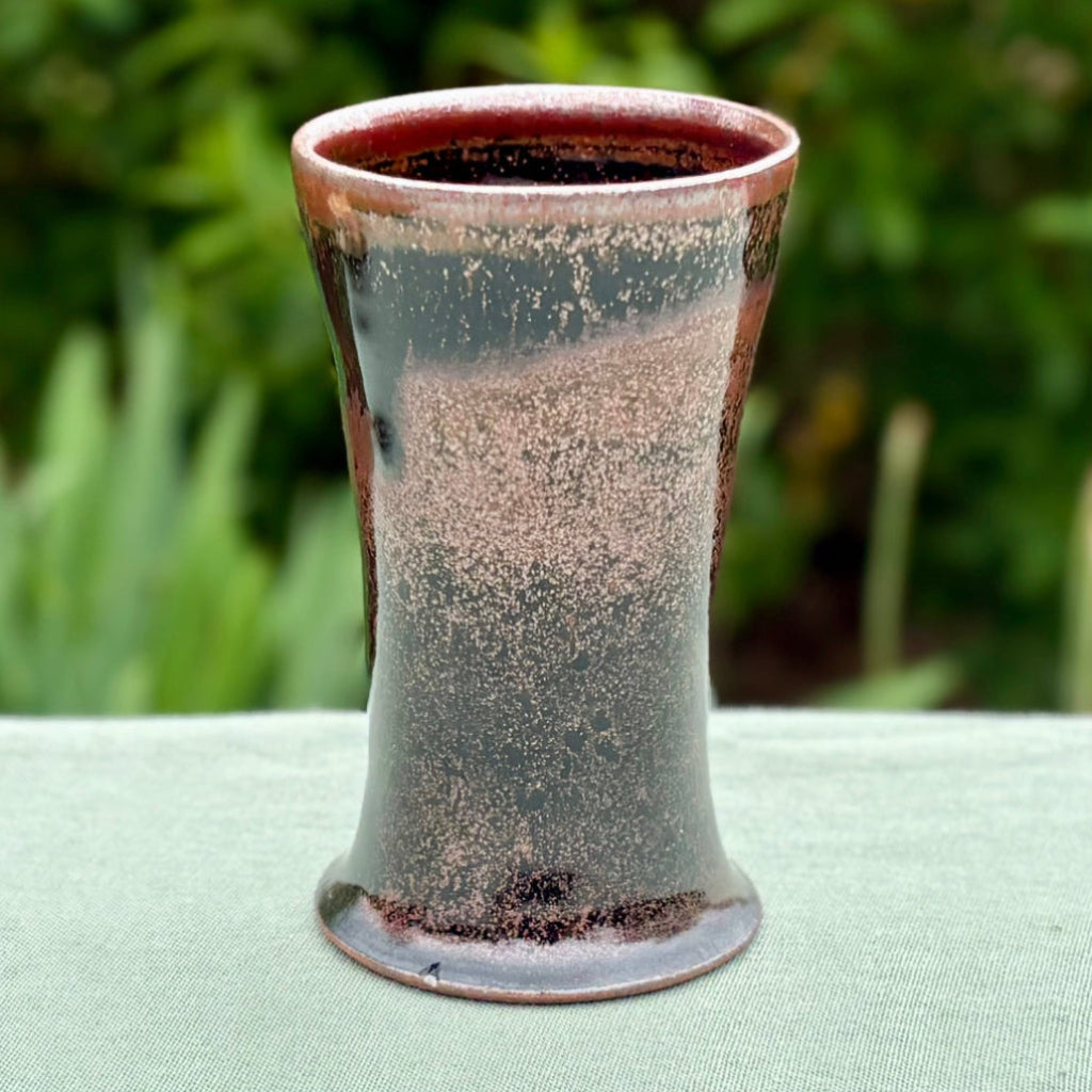Handcrafted wood-fired cup with red/pink speckles and flared rim on countertop with herbs next to the cup. 