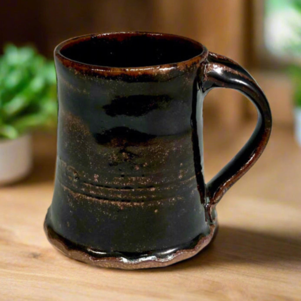 Handcrafted rustic black Tenmoku stoneware mug with brown rim accent and curved handle on a counter top with window behind it and flowers, wood-fired in Danbury, NC.