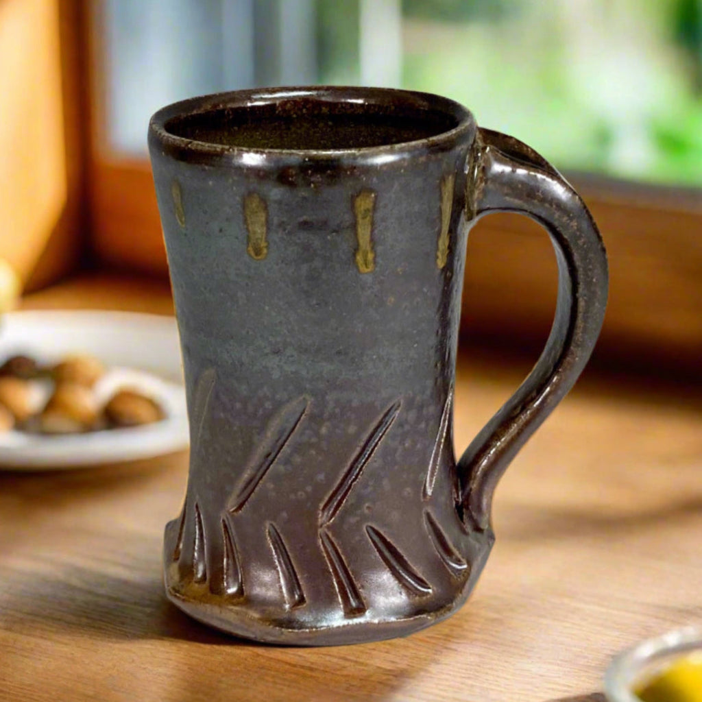 Handcrafted gray stoneware mug with carved chevron pattern, amber glaze drips and curved handle on a counter top with window behind it and a place of food.