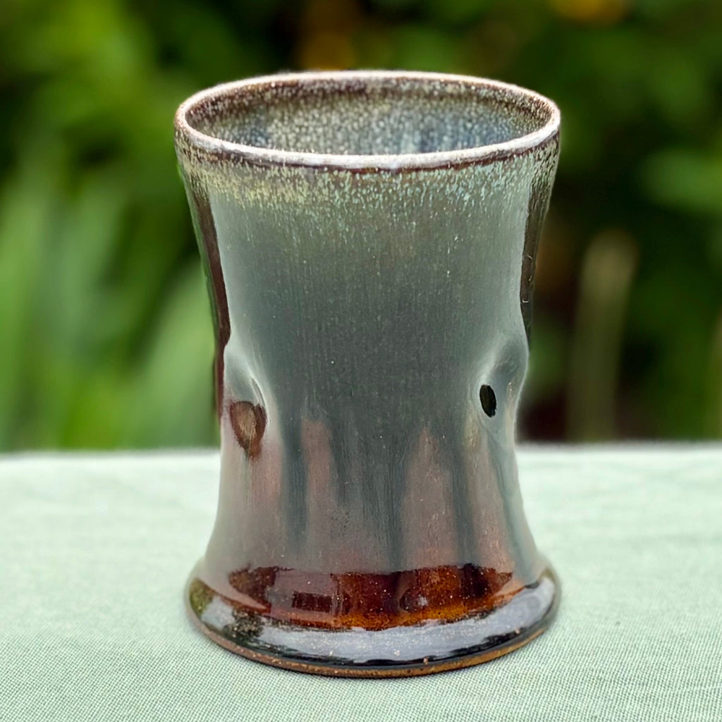 Handcrafted wood-fired ceramic cup with moss green interior transitioning to rich amber-brown base on green tabletop with greenery in the background. 