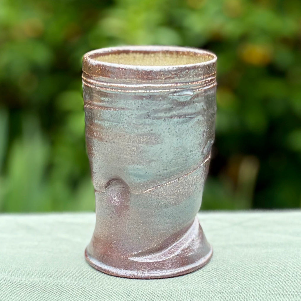 handcrafted wood-fired pottery cup with green-dark brown exterior on green fabric tabletop with greenery in the background. 