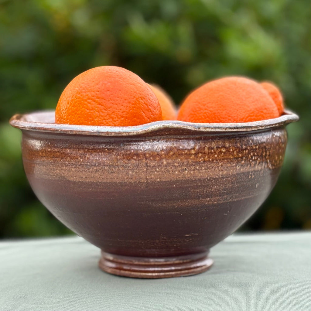 Handcrafted amber and brown speckled stoneware serving bowl with smooth interior and textured gradient exterior with greenery in background. 