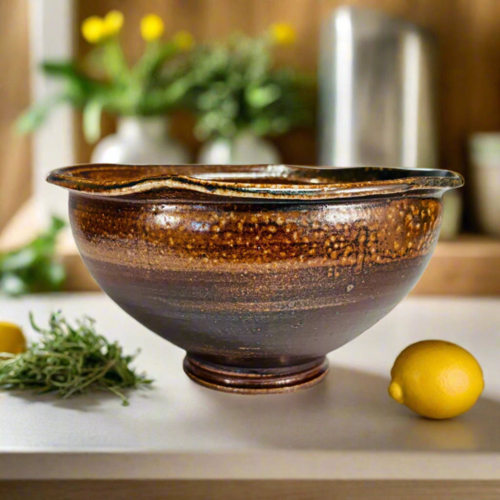 Handcrafted amber and brown speckled stoneware serving bowl with smooth interior and textured gradient exterior with citrus and herb and flowers in the background.