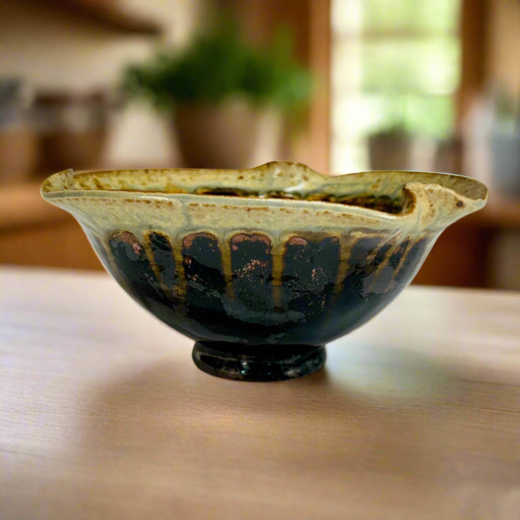 A handcrafted ceramic bowl with a two-tone glaze, deep black base transitioning to speckled beige at the rim, with an asymmetrical curved edge.