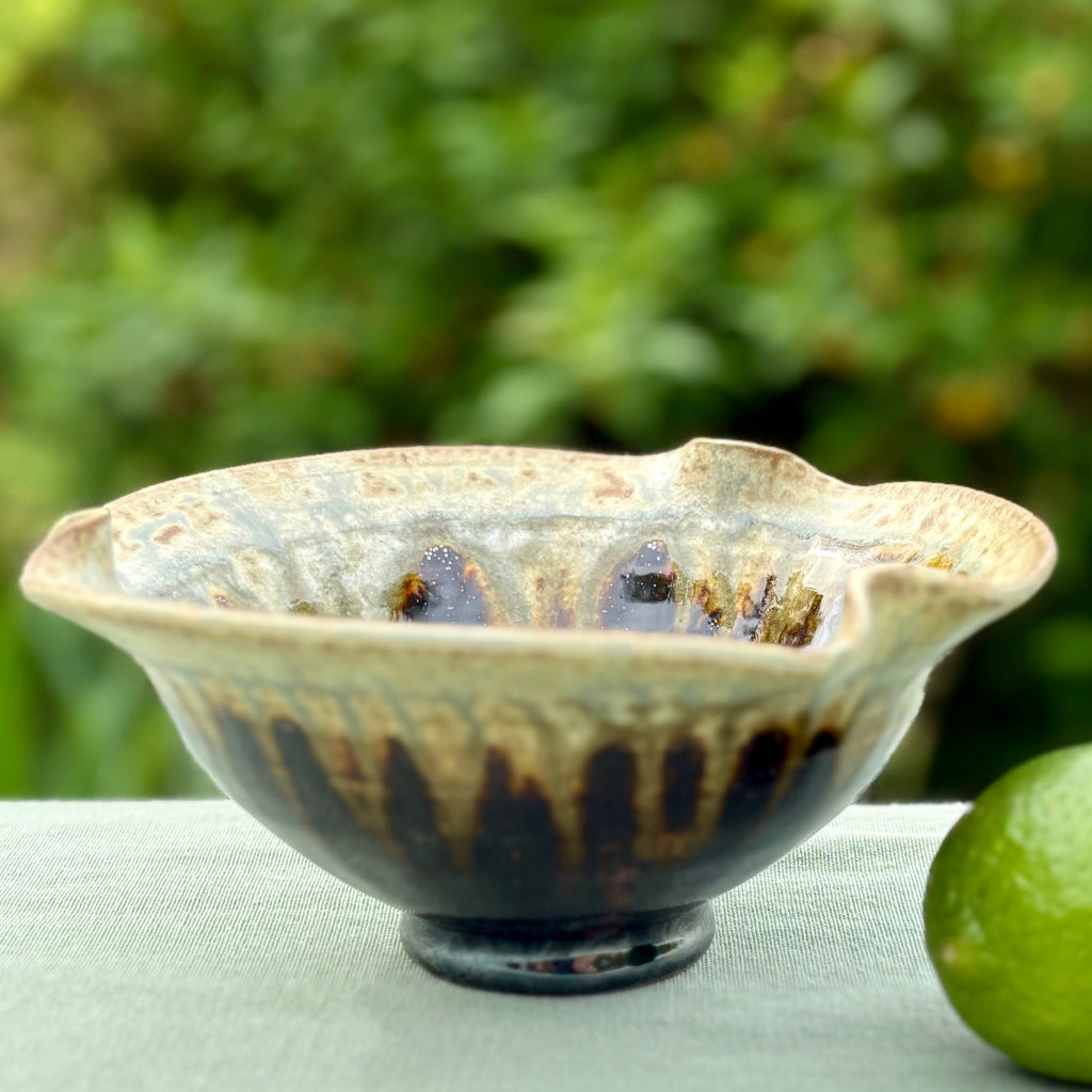 A handcrafted ceramic bowl with a two-tone glaze, featuring a deep black base and a blue ash rim, displayed outdoors with a green blurred background and a lime to the side.