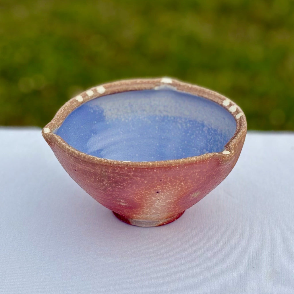 Handmade porcelain bowl with blue interior and red-brown exterior on a white surface with a blurred green background