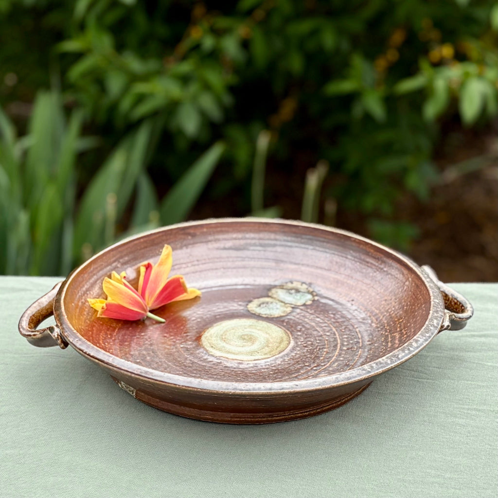 A ceramic platter with a wood flame and salt glaze, featuring a white glaze in the center and two small handles. The platter is displayed on a table with a natural background and a small flower.