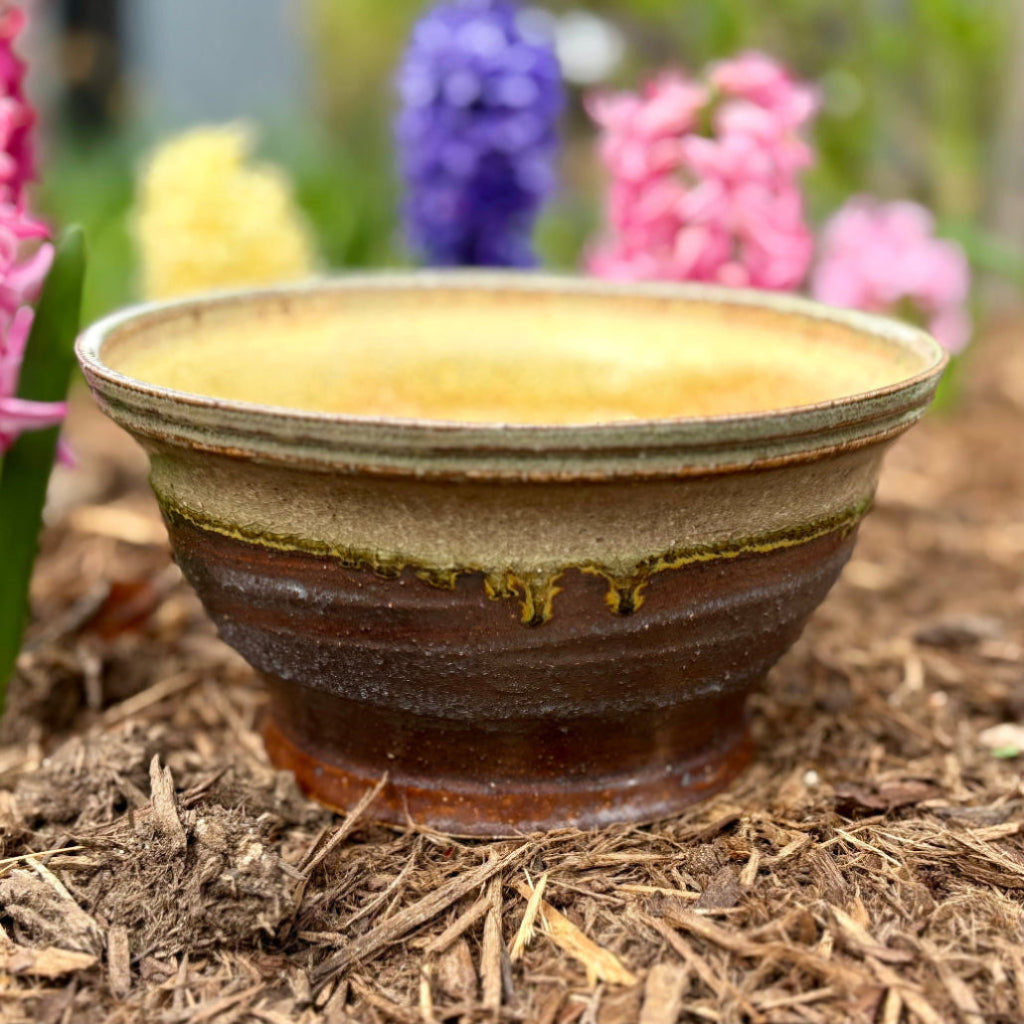 Handcrafted blue-gray and cream ceramic planter with drainage hole, side profile showing textured glaze with flowers in the background. 