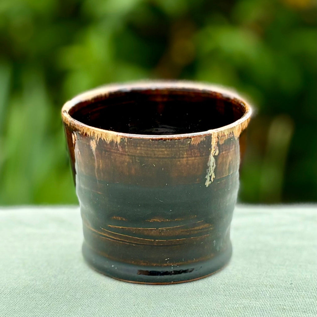 small ceramic cup with brown and black glazes and a unique 'drip' effect along the rim, sitting on a green surface with foliage in the background