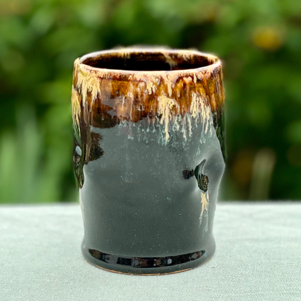 wheel-thrown ceramic cup with a deep ebony gloss tenmoku glaze and golden-amber drips on a light-colored surface with foliage in the background