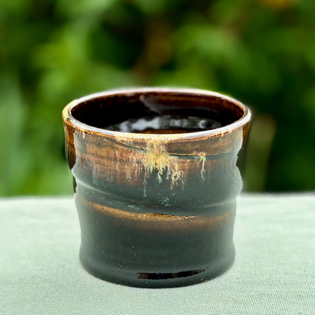 Handcrafted tenmoku ceramic cup with rich black glaze and amber gold drip detail, featuring slight asymmetrical form on green cloth with greenery in the background. 