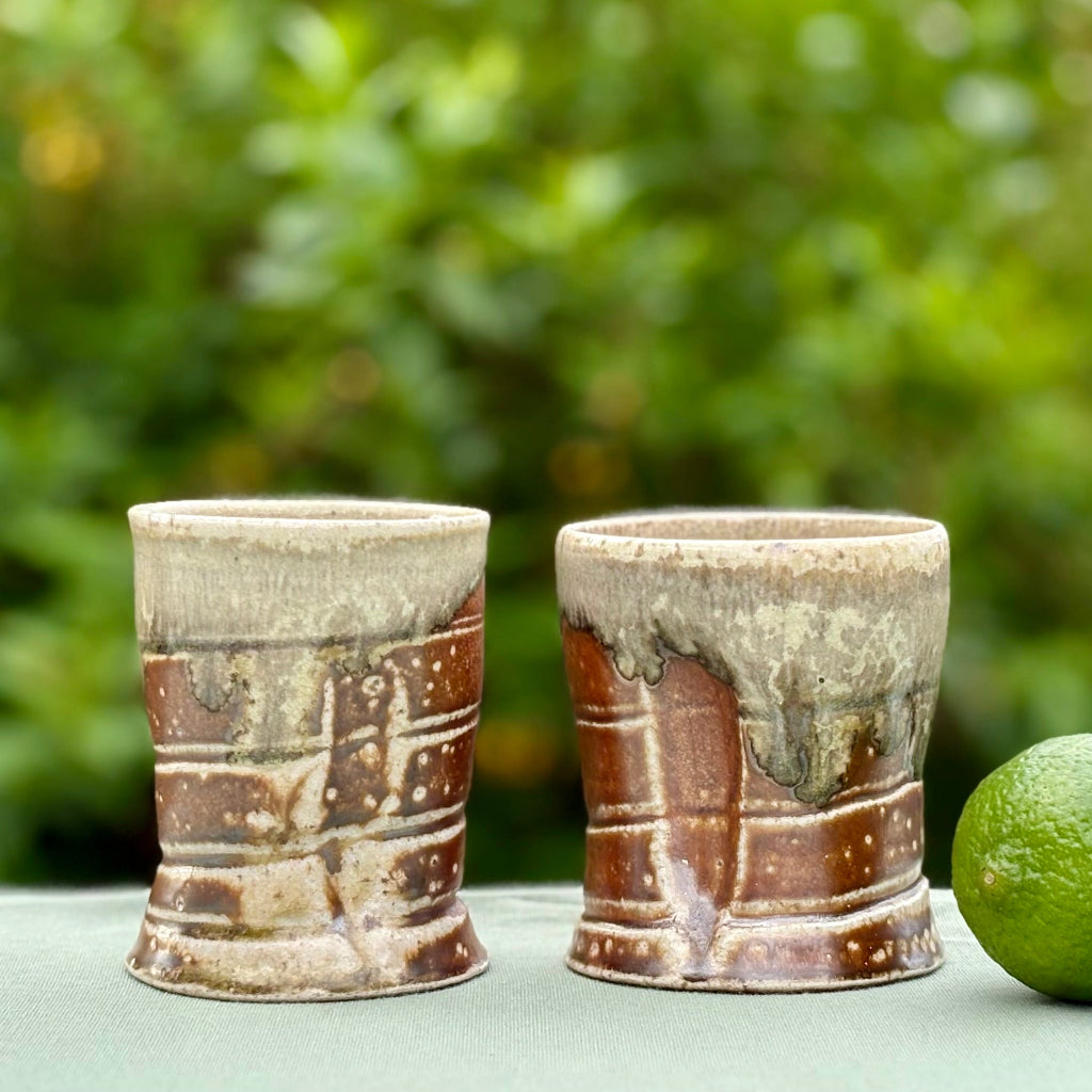 two Harmony Stoneware Cups featuring a celadon glaze on the inside and orange flashing slip on the outside, placed on a table with a green background