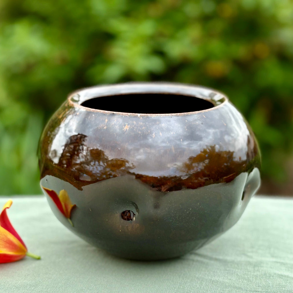 Round chestnut glazed vase with a dimpled surface on a green cloth with a flower and greenery in the background, wood-fired ceramics in Danbury, NC