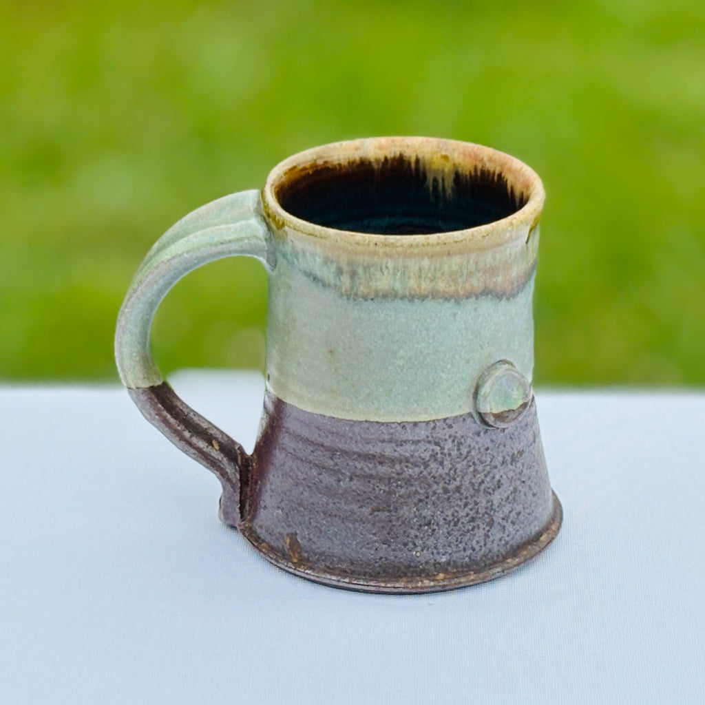 Oribe ceramic mug with a deep brown/black interior, an unglazed, earthy lower half and a variegated green upper half. A small button adds visual and tactile interest