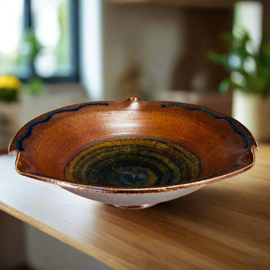Handcrafted ceramic serving bowl with wavy rippled edge, blue accent lines, and warm brown glaze with golden-green center on wooden counter top with plants in the background.
