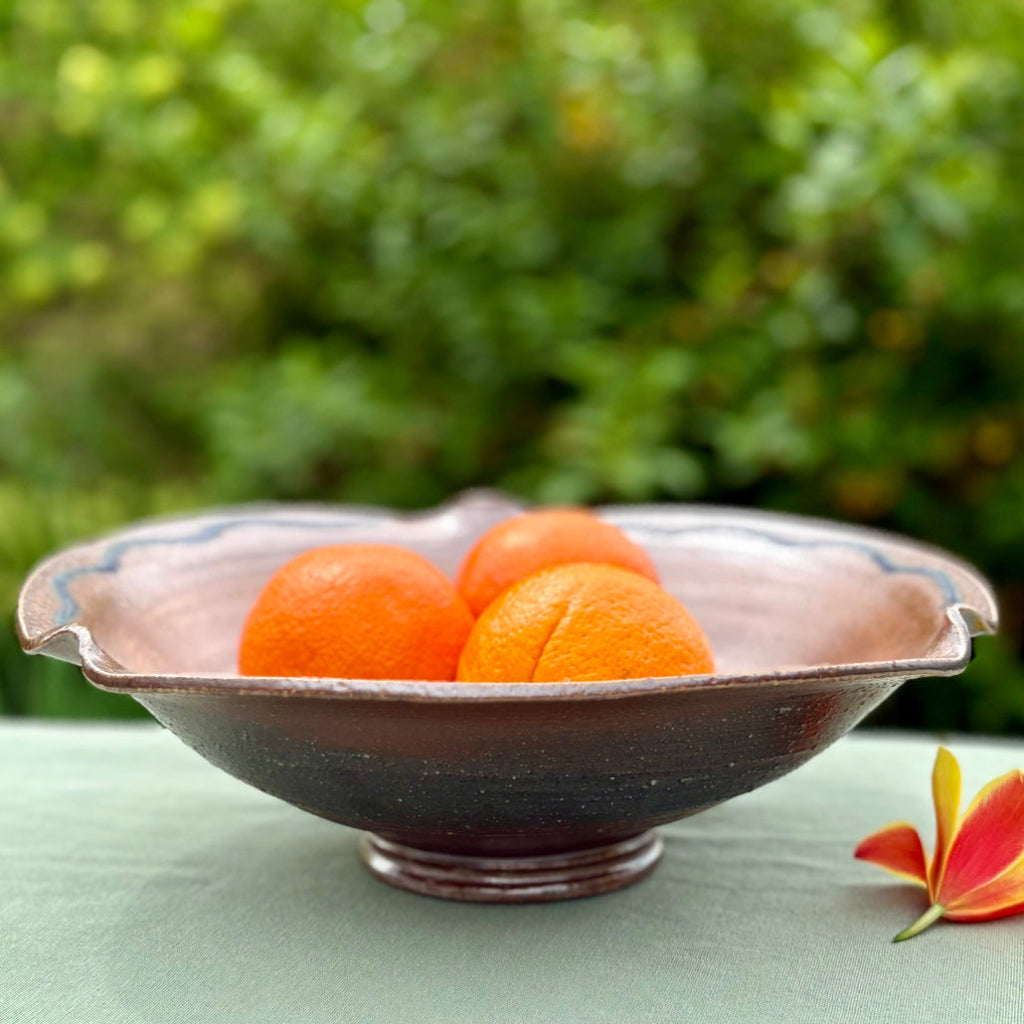 Handcrafted ceramic serving bowl with wavy rippled edge, blue accent lines, and warm brown glaze with golden-green center on green cloth with greenery in the background.