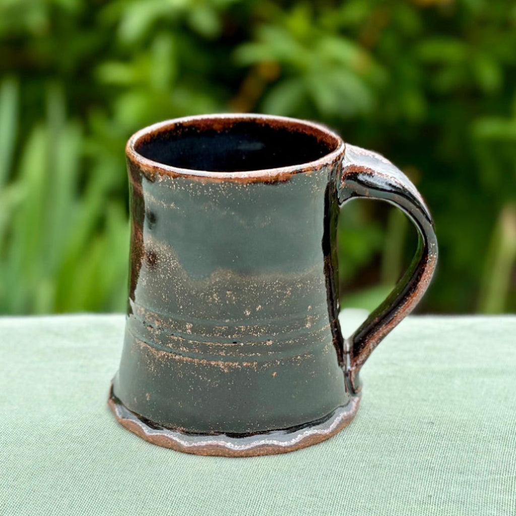 Handcrafted rustic black Tenmoku stoneware mug with brown rim accent and curved handle on a green cloth with greenery in the background, wood-fired in Danbury, NC.
