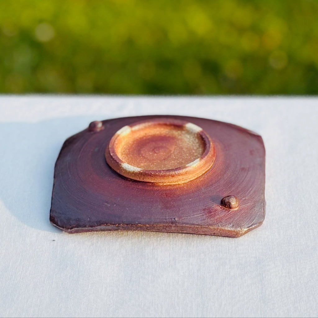 Square ceramic soap dish with a deep reddish brown bottom and a circular base with four white dots, a faint swirl and two small round feet 