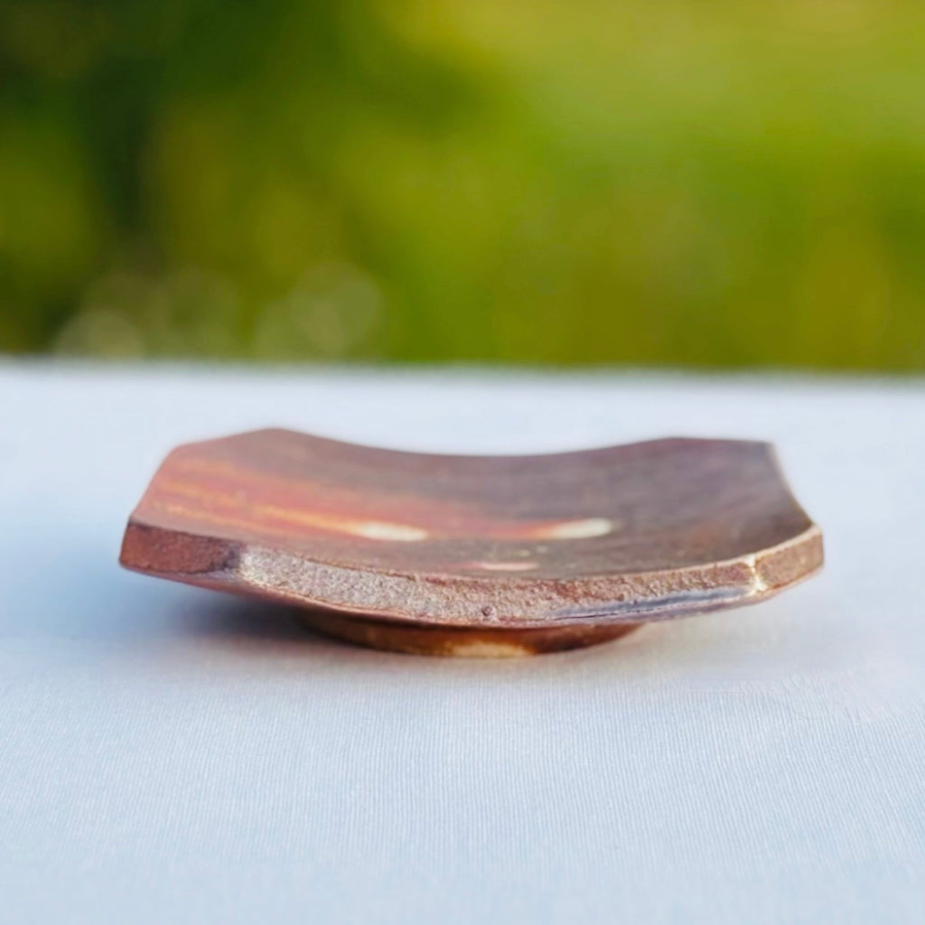 Wood-fired ceramic soap dish with natural ash glaze showing orange flame patterns and earth tones with three white dots in the center and a subtle lines
