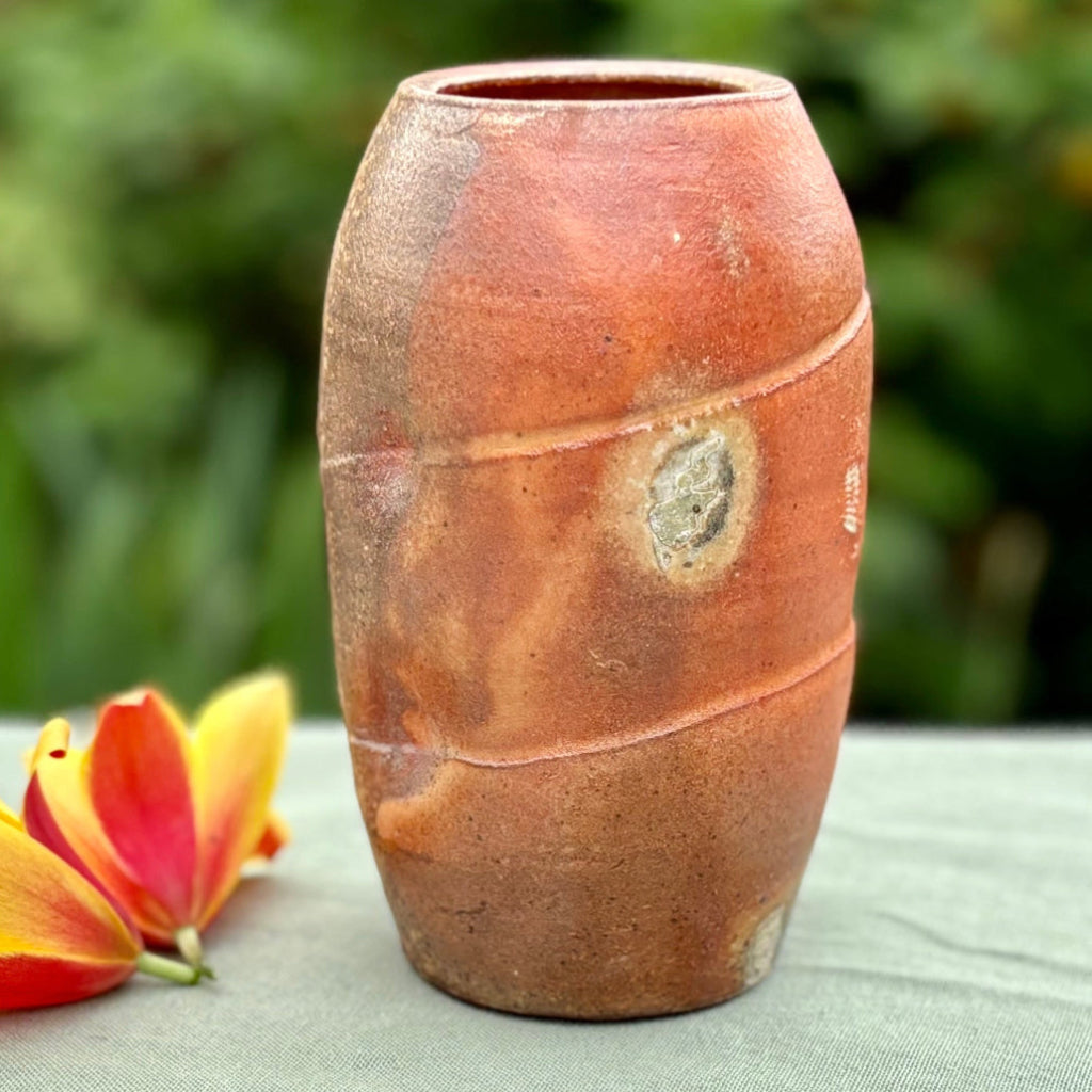 Handcrafted wood-fired ceramic vase in vibrant orange with natural ash deposits and horizontal banding on green cloth with a flower with greenery in the background