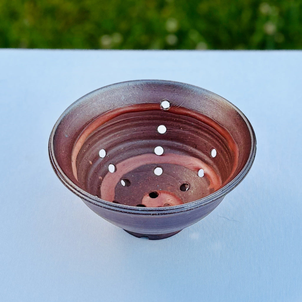 Unglazed handmade stoneware bowl showing wood kiln fire effects in rich reddish browns and amber spiral pattern and drain holes 