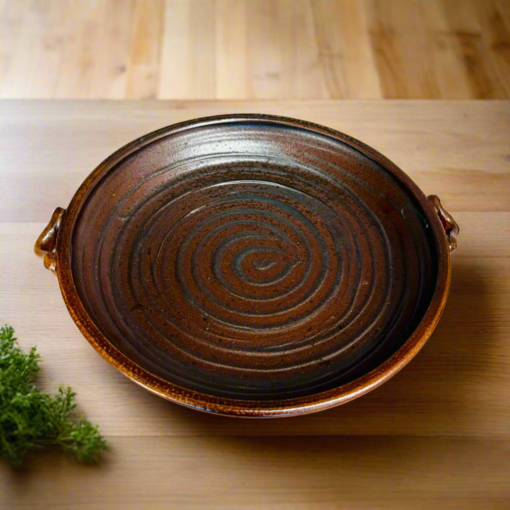 Handcrafted ceramic serving platter with blue spiral design on mahogany brown background and integrated handles on countertop with herbs next to the dish.