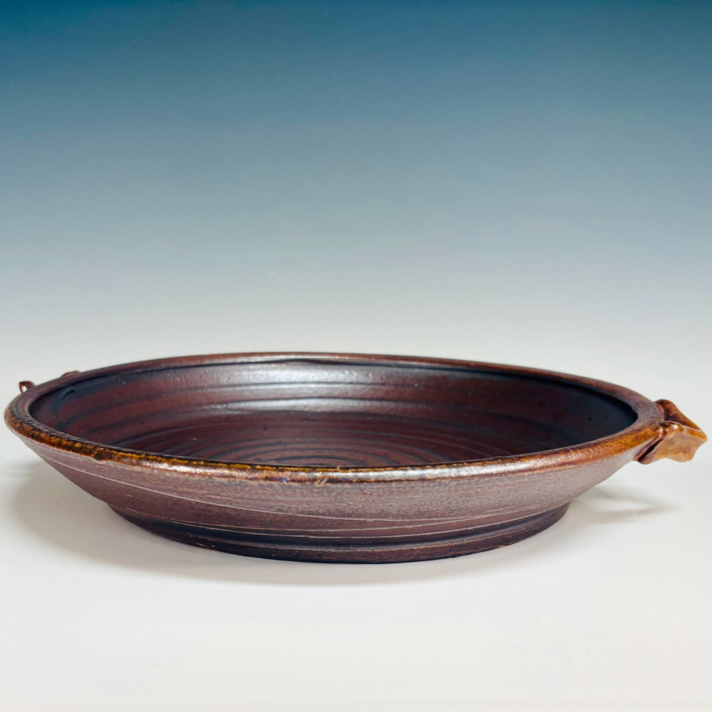 Side view: Handcrafted ceramic serving platter with blue spiral design on mahogany brown background and integrated handles