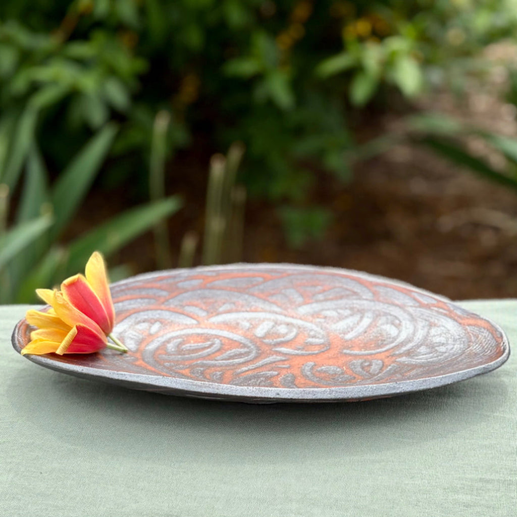 Decorative oval ceramic serving tray with delicate brown-black swirls over an orange flashing base on a green cloth with green in the background