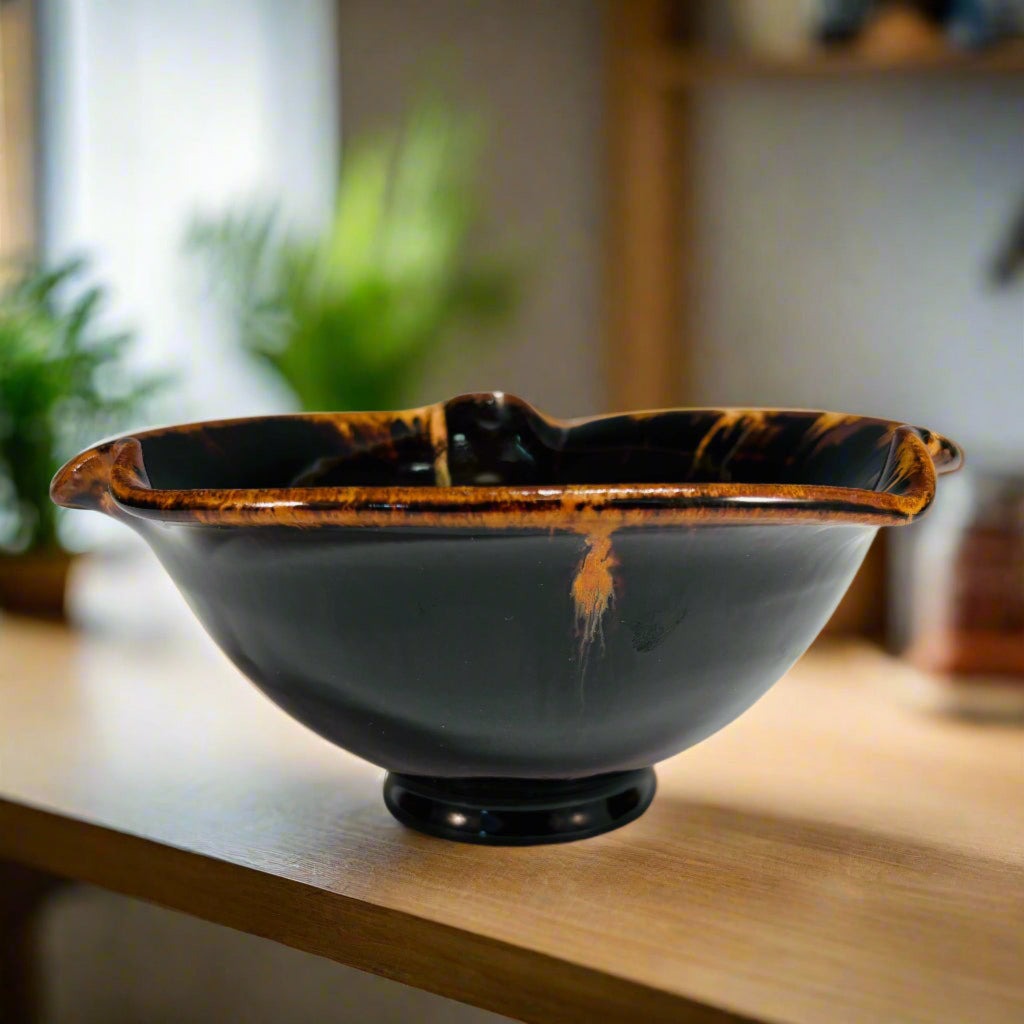 dark brown/black pottery bowl with high shine glaze and a amber rims and drips on a wooden kitchen countertop 